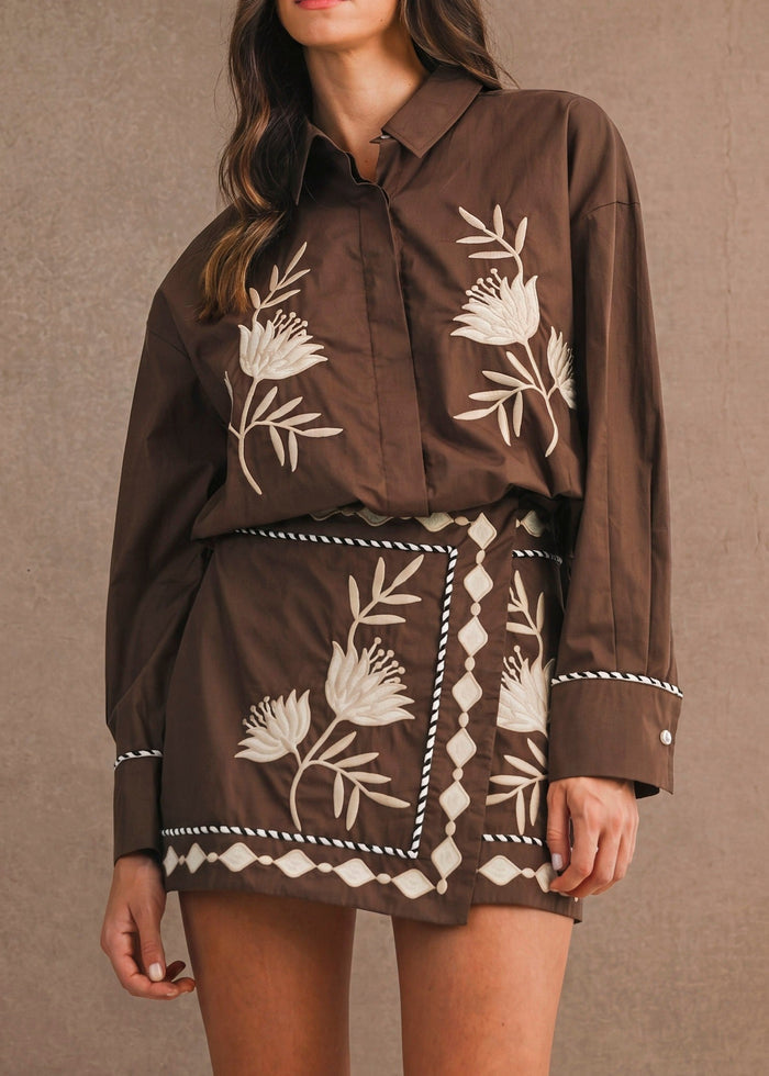 A woman modeling a brown button-up shirt and matching mini skirt set with white floral and geometric embroidery, set against a textured brown studio backdrop.