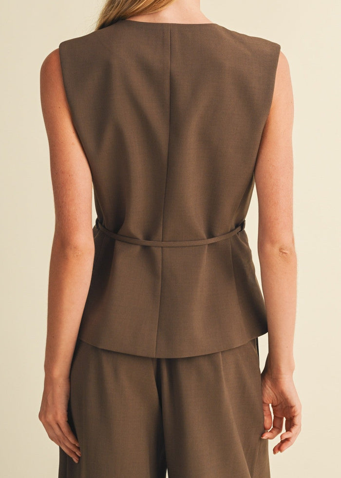 A woman shown from the back wearing a sleeveless brown peplum-style top and matching brown trousers against a plain warm beige studio background.