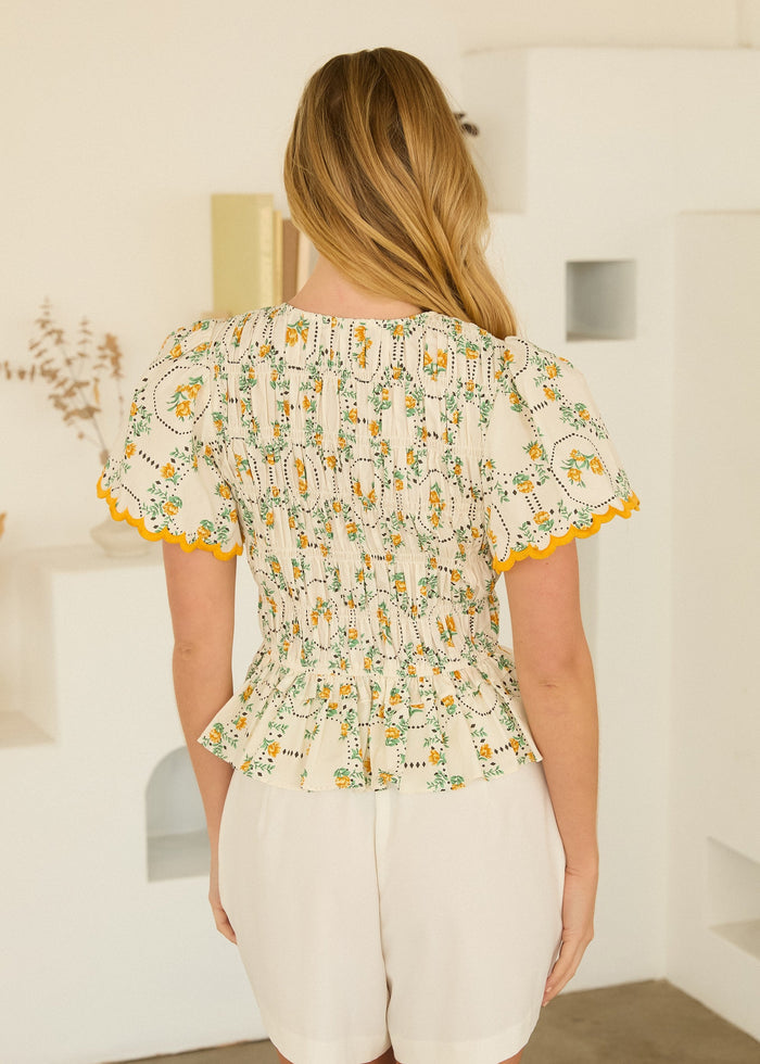 Cream peplum top with yellow and green floral print, scalloped orange trim, and short flutter sleeves.