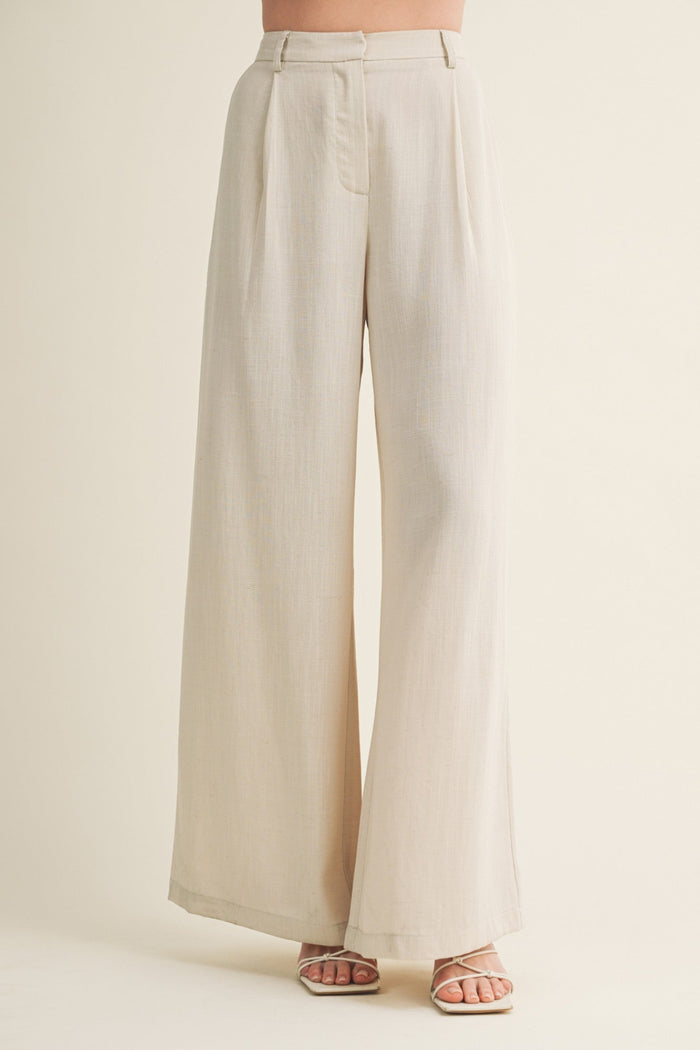 Cream wide-leg pants with pleated front and high waistband on model wearing strappy sandals.
