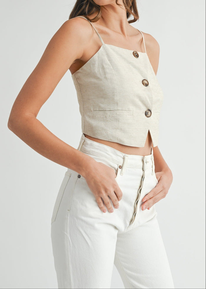 Beige linen crop top with button front and spaghetti straps worn with white high-waisted pants.