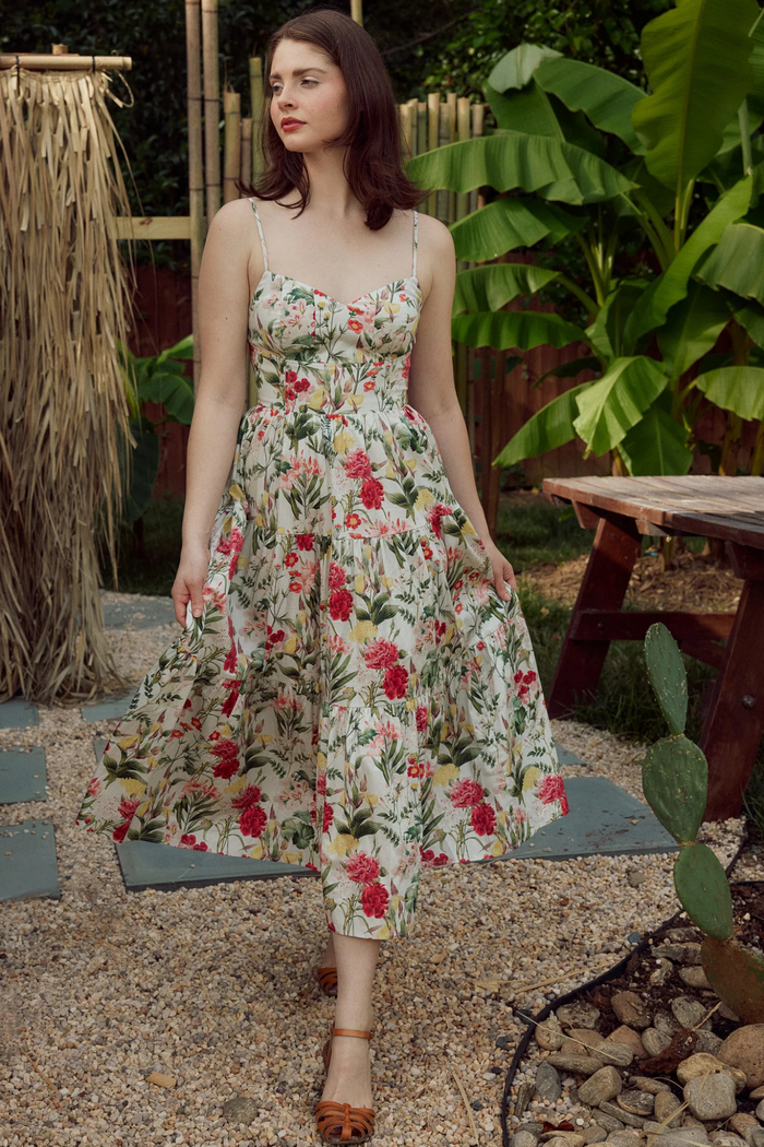 Woman wearing cream maxi dress with red and pink floral print, spaghetti straps, and fitted bodice in garden setting.