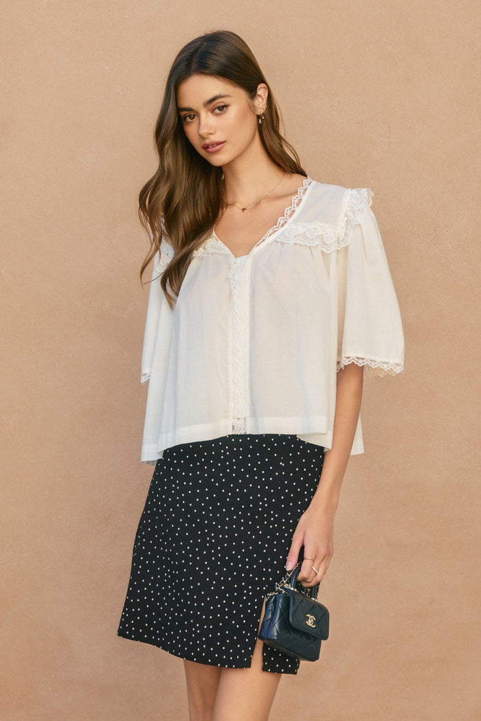 White lace shirred blouse with V-neckline and ruffled collar, displayed on model with black polka dot skirt.