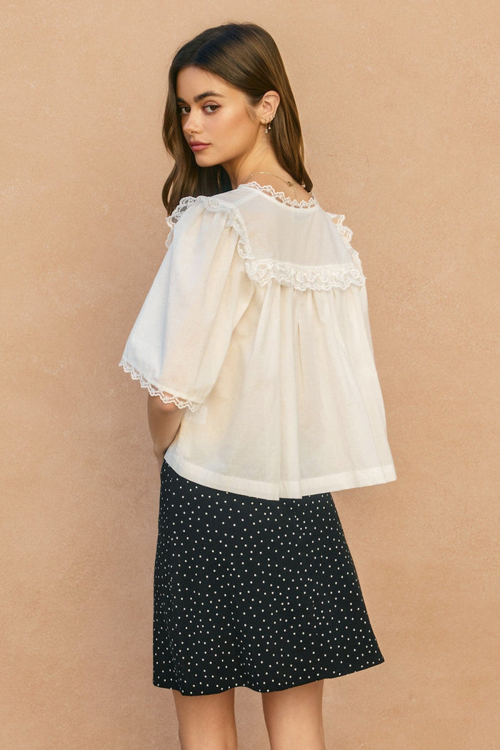 Cream lace-trimmed shirred blouse with ruffled cape sleeves worn over black polka dot skirt.
