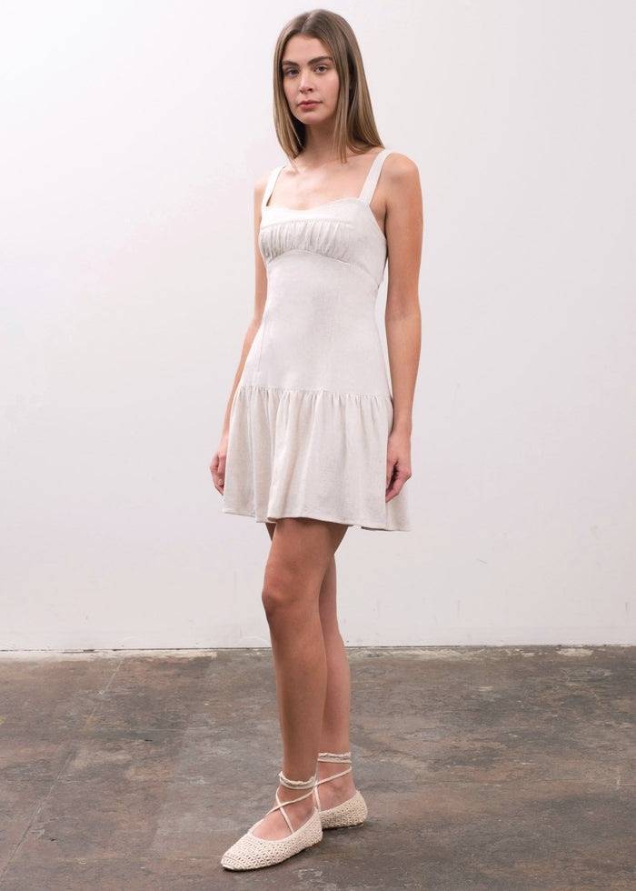 Sleeveless white cotton dress with square neckline, ruched detail at bust, and gathered skirt hem on model wearing white stra
