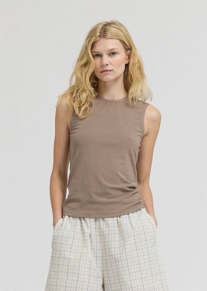 Taupe sleeveless tank top with wide shoulders and crew neckline on model wearing cream textured pants.