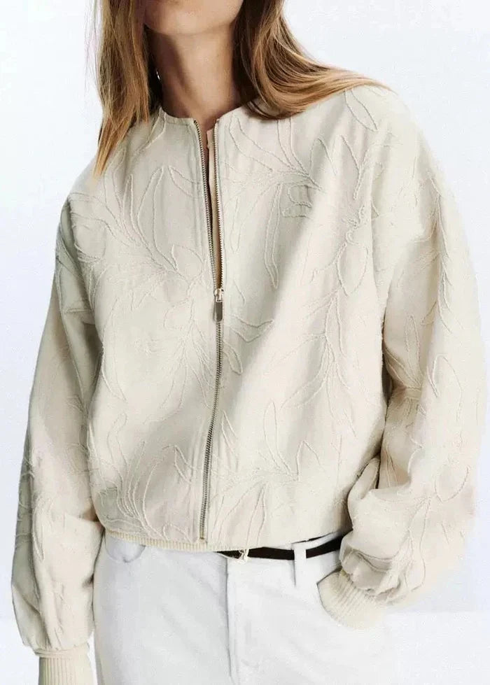 Cream quilted zip-up jacket with leaf pattern and ribbed cuffs, worn over white pants.