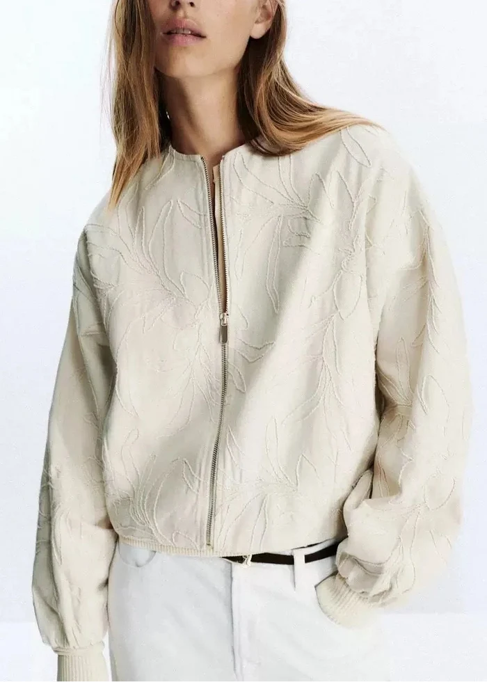 Cream quilted zip-up jacket with leaf embroidery pattern and ribbed cuffs worn with white pants.