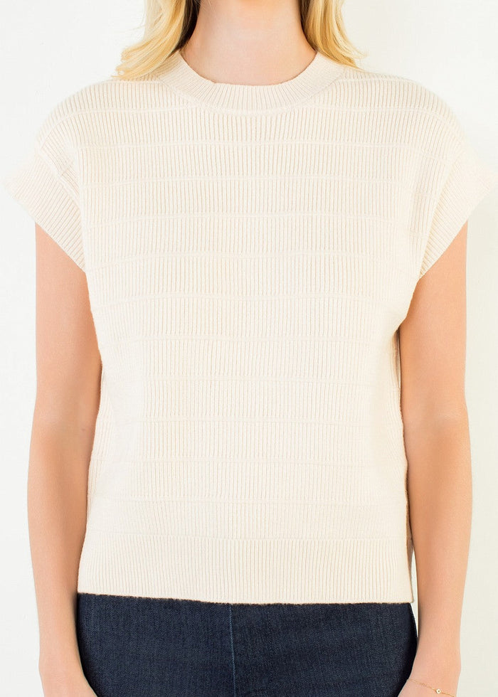 A woman wearing a cream-colored ribbed short-sleeve knit top and dark blue jeans, shown from the neck down against a plain white background.