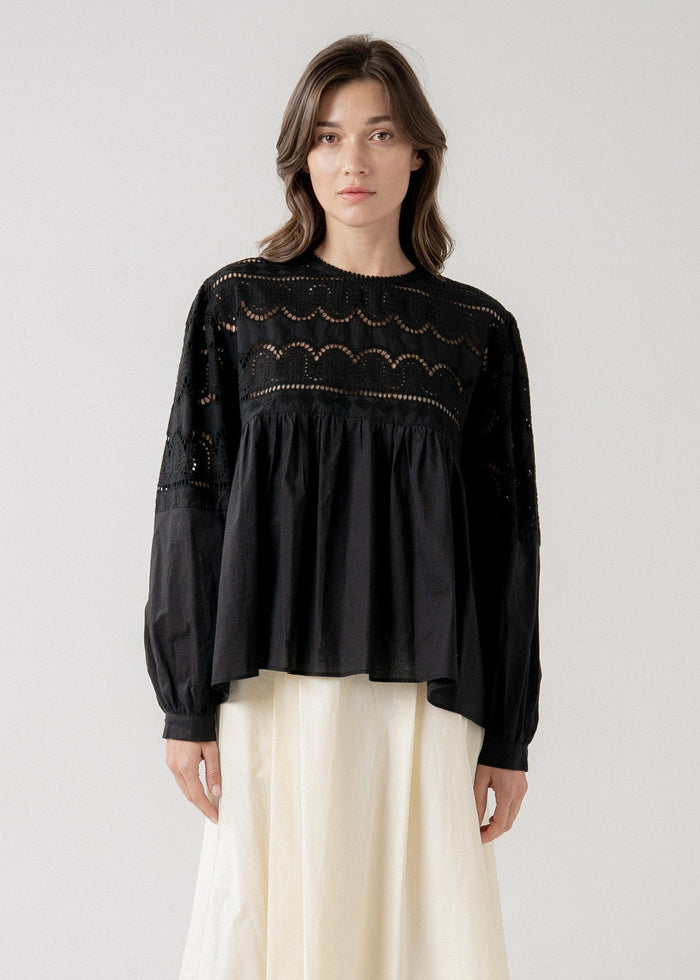 Black long-sleeve babydoll blouse with lace yoke and gathered waist over cream skirt.