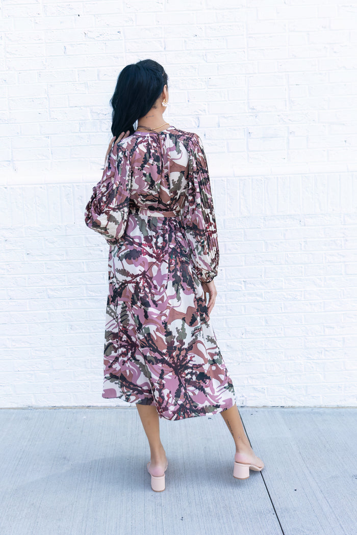 Woman wearing midi dress with purple and brown abstract floral print, long puffed sleeves, and wrap-style waist, shown from b