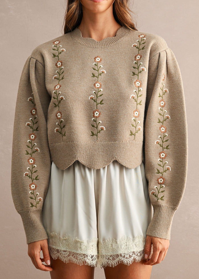 A woman wearing a taupe knit sweater with vertical floral embroidery down the front and along the sleeves, paired with light sage green shorts, against a neutral beige background.