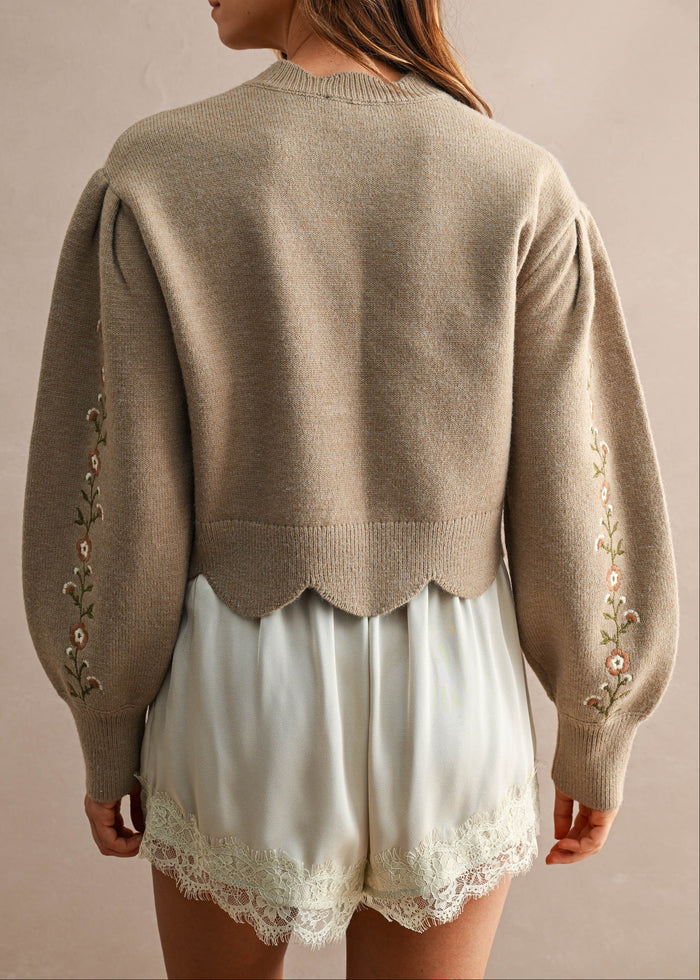 A woman shown from the back wearing a taupe knit sweater with decorative floral embroidery down the sleeves and a scalloped hem, paired with white lace-trimmed shorts against a neutral beige background.