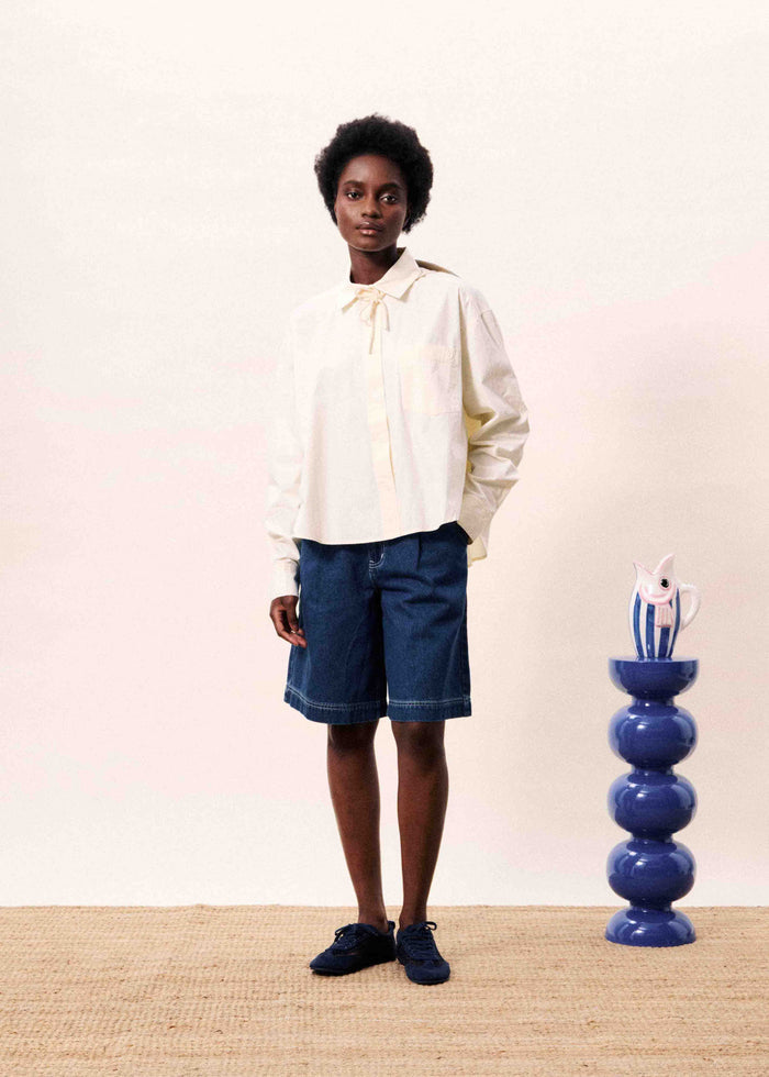 Cream button-down shirt with tie neckline worn with denim shorts.
