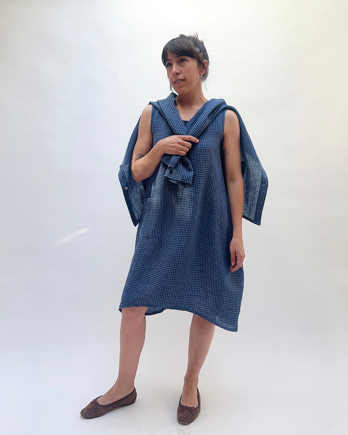 Christopher Calvin Linen Mini Check Dress in navy-and-white mini check, worn with a matching linen shirt draped over the shoulders, showing the scoop neckline, sleeveless silhouette, and asymmetric hem, with brown ballet flats on a white background