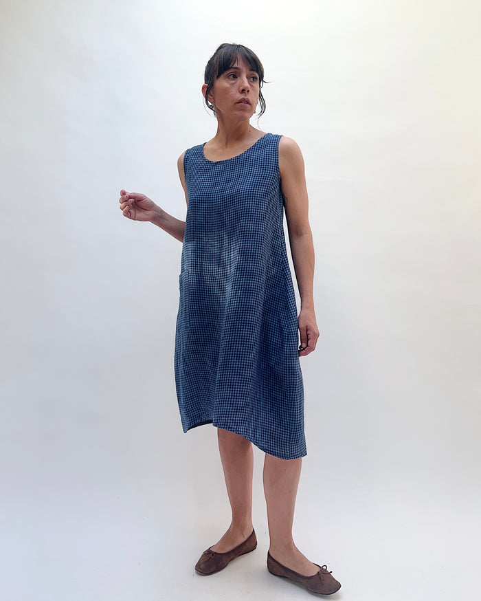 Three-quarter front view of Christopher Calvin Linen Mini Check Dress in navy-and-white mini check, showing the sleeveless cut, relaxed drape, and asymmetric hem falling below the knee, worn with brown ballet flats on a white background