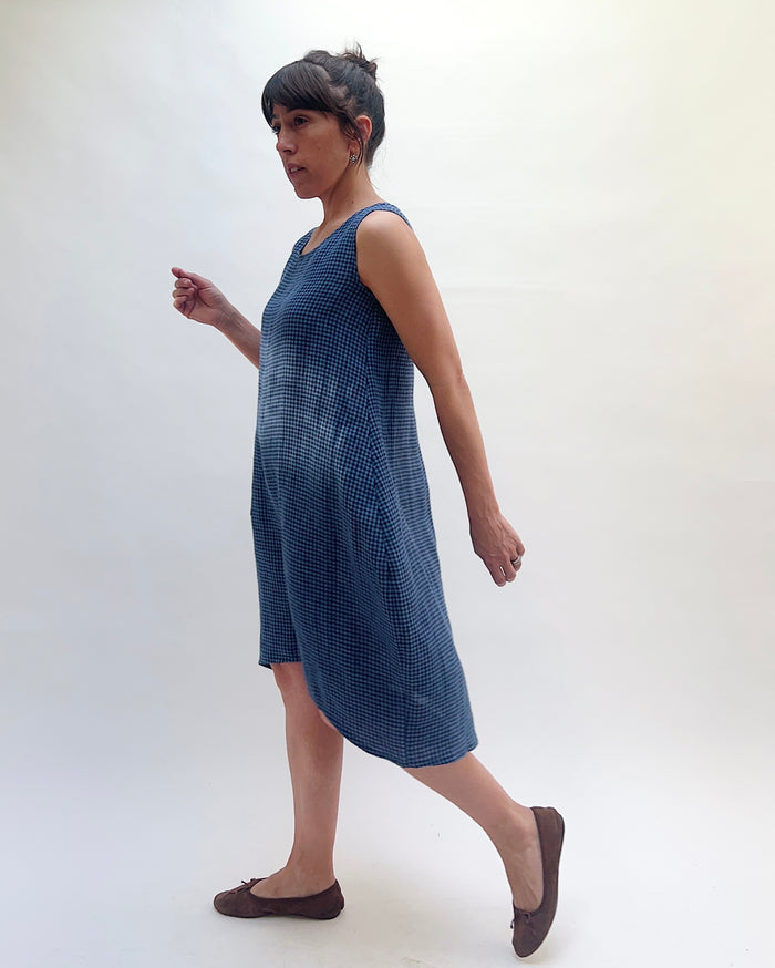 Side profile of Christopher Calvin Linen Mini Check Dress in navy-and-white mini check, showing the scoop neckline, sleeveless silhouette, and the longer back hem of the asymmetric cut, worn with brown ballet flats on a white background