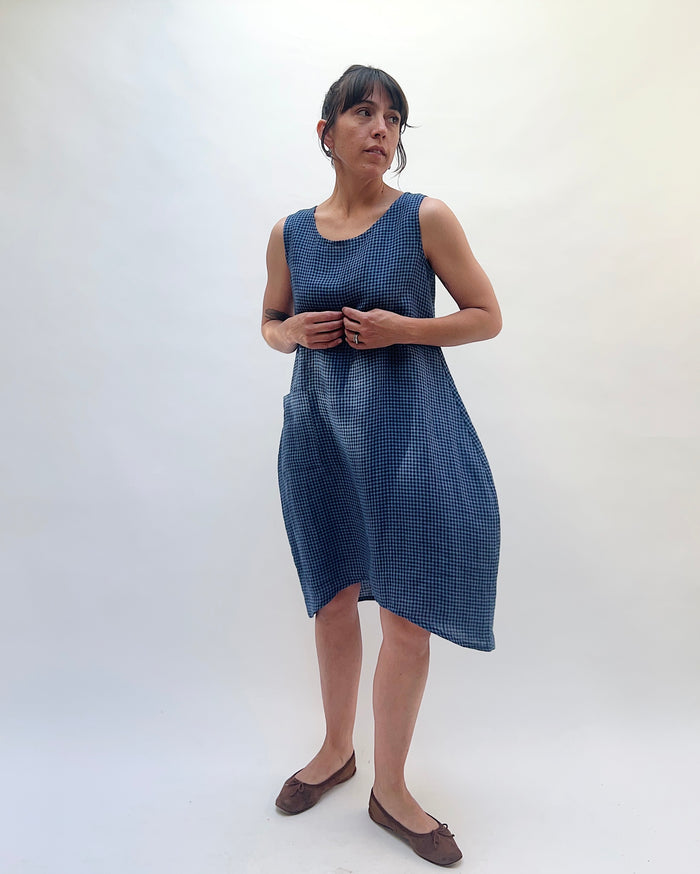 Front view of Christopher Calvin Linen Mini Check Dress in navy-and-white mini check with hands gathered at the waist, showing the relaxed boxy drape, side pocket, and asymmetric hem, worn with brown ballet flats on a white background
