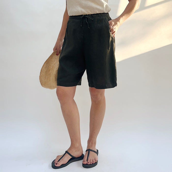 Three-quarter front view of model wearing Bryn Walker peppercorn linen sasha short, showing elastic drawstring waistband, side pocket, relaxed wide-leg bermuda length, and deep charcoal color, with straw hat and black thong sandals
