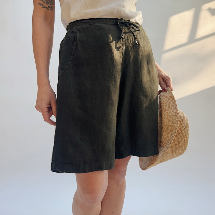 Close three-quarter front view of model wearing Bryn Walker peppercorn linen sasha short, showing elastic drawstring waistband, side pocket, relaxed wide-leg bermuda length, and deep charcoal color, with straw ha