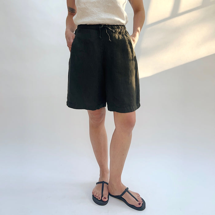 Full front view of model wearing Bryn Walker peppercorn linen sasha short with hands in pockets, showing elastic drawstring waistband, relaxed wide-leg bermuda length, and deep charcoal color, with black thong sandals