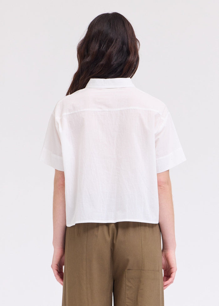 White short-sleeve button-up top with collar, shown from back view on model wearing tan pants.