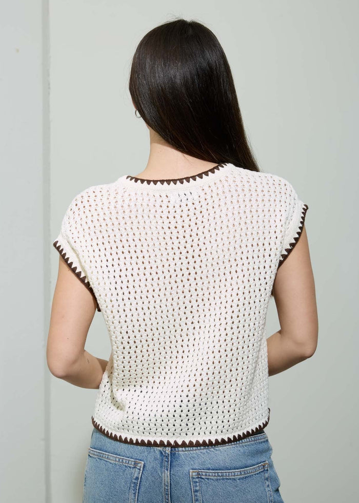 Woman wearing a white crochet top with brown trim and blue jeans against a plain background