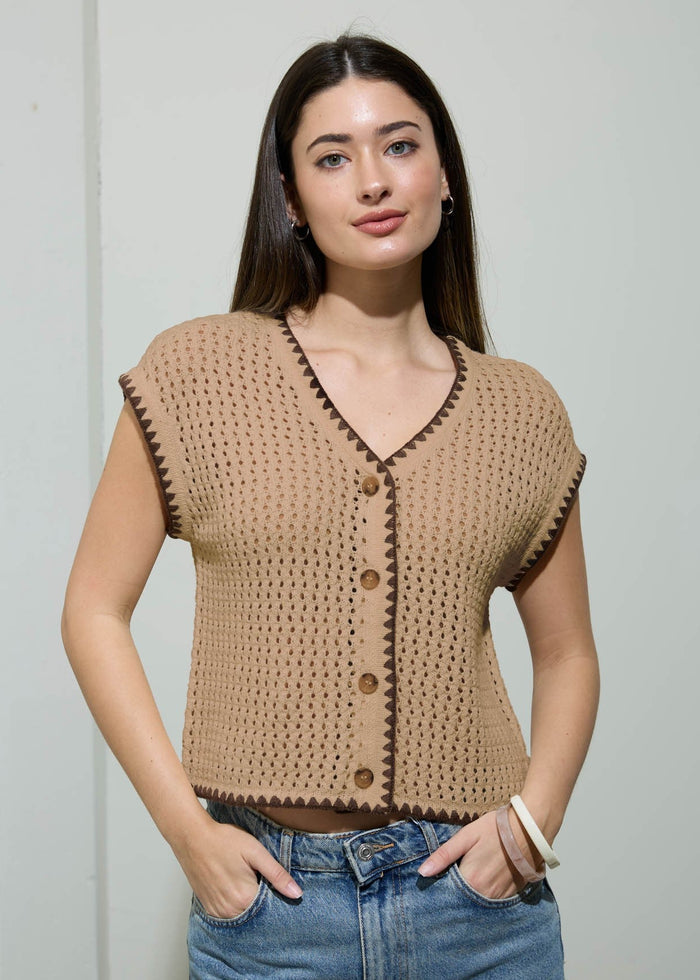 Woman wearing a beige knitted vest over a white shirt with blue jeans against a plain background