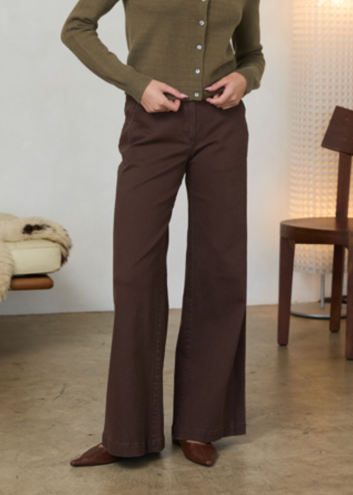 A person wearing dark brown wide-leg pants and an olive green button-front top, standing in a softly lit indoor room with a chair and textured wall visible in the background.