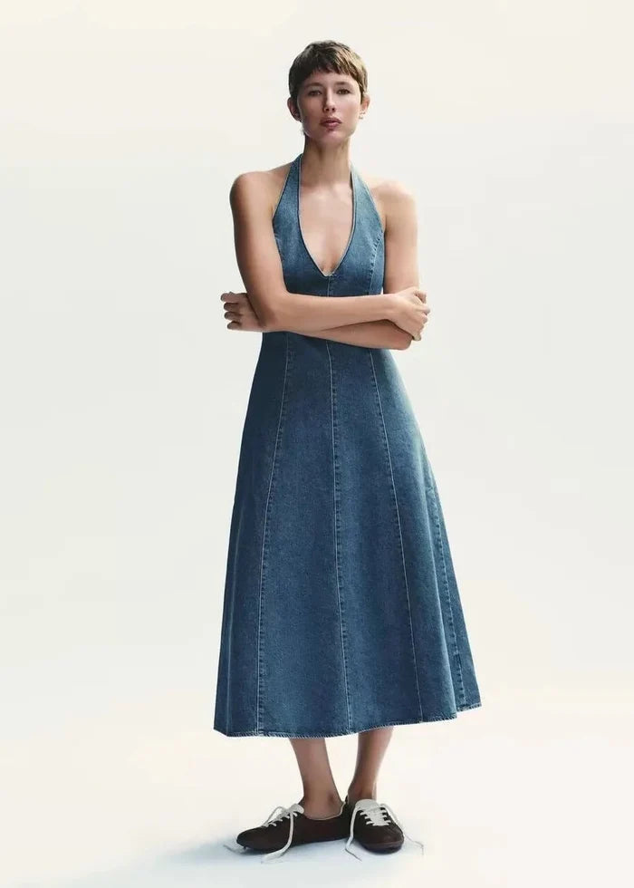 Blue denim halter maxi dress with a V-neckline and A-line skirt on a model wearing black and white sneakers.