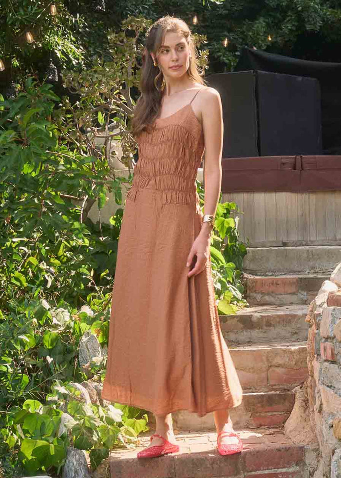 Woman wearing a rust-brown sleeveless midi dress with shirred bodice and waist, standing on brick steps.