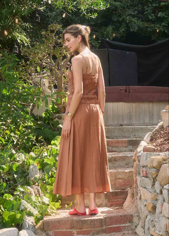 Woman wearing a rust-brown sleeveless midi dress with shirred detailing, standing in a garden setting.