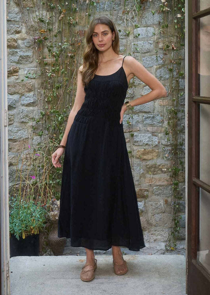 Black sleeveless midi dress with shirred bodice and thin straps on model against stone wall.