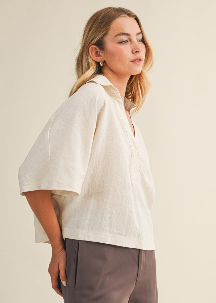 Woman wearing cream linen popover shirt with collar and short sleeves, paired with taupe trousers.
