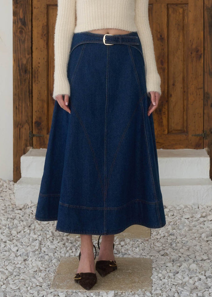 Woman wearing a white sweater and blue skirt standing in front of a wooden door.