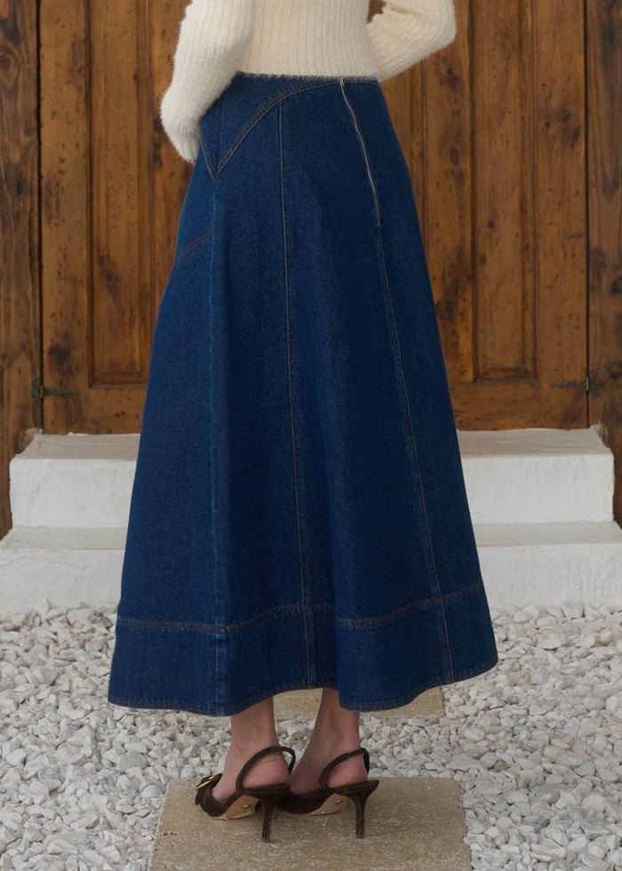 Woman in a white sweater and blue skirt standing in front of a wooden door.