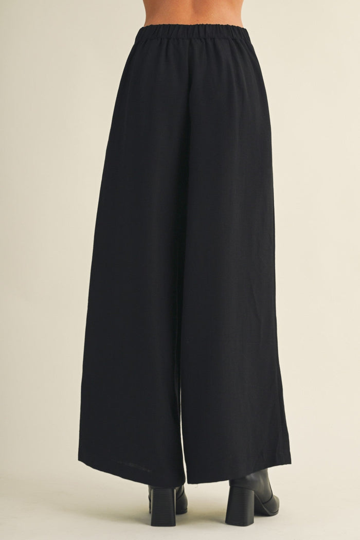 Black wide-leg cropped pants shown from the back on a model, featuring an elastic waistband and a straight, flowing silhouette, paired with black block-heeled boots against a light beige background.