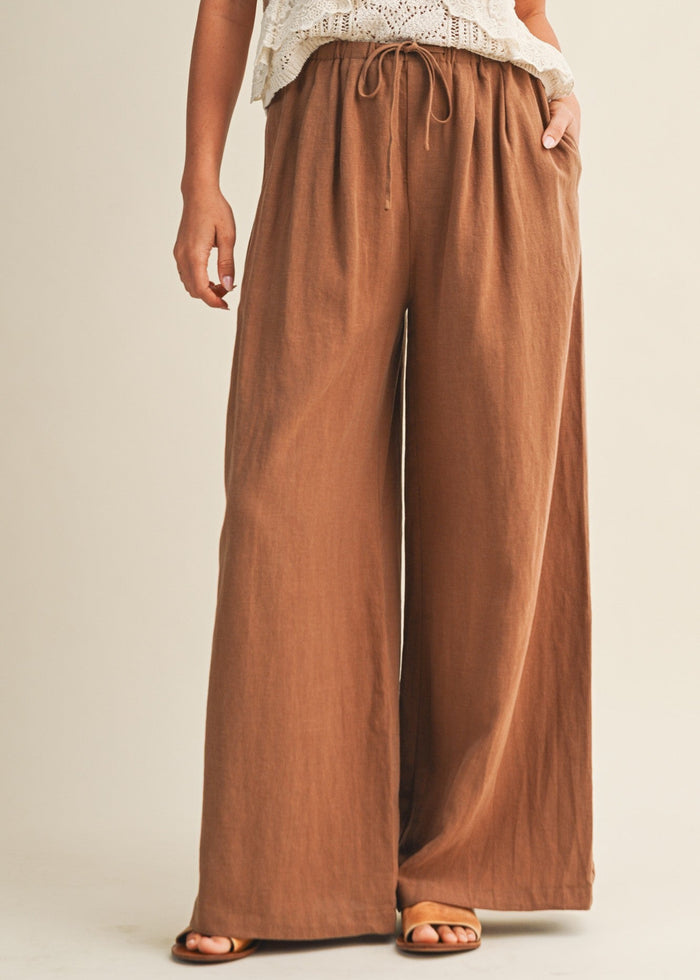 Tan wide-leg pants with drawstring waist and side pockets worn with white crochet top and tan slides.