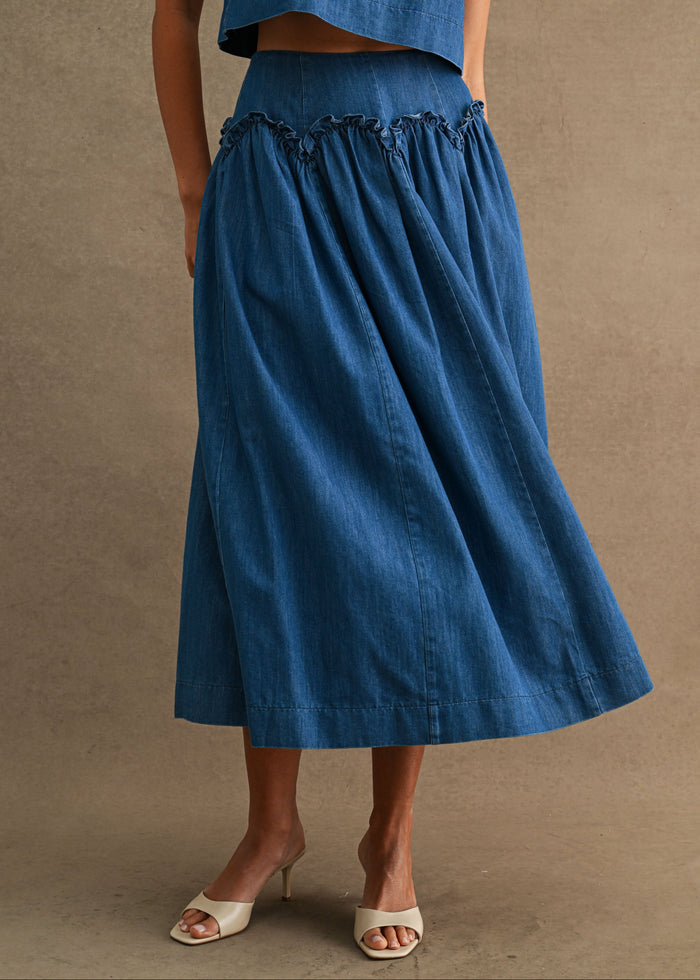A model wearing a blue denim midi skirt with a ruffled waistband and a matching blue denim cropped top, styled with white heeled sandals against a textured beige background.