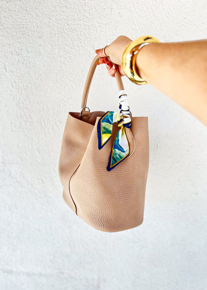 Beige bucket tote handbag with tan handles and blue, yellow, and green patterned scarf tied to the handle.