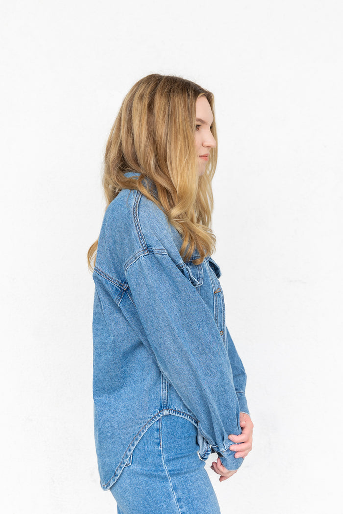 Blue denim button-up shirt with chest pockets and curved hem, shown on model in profile view.