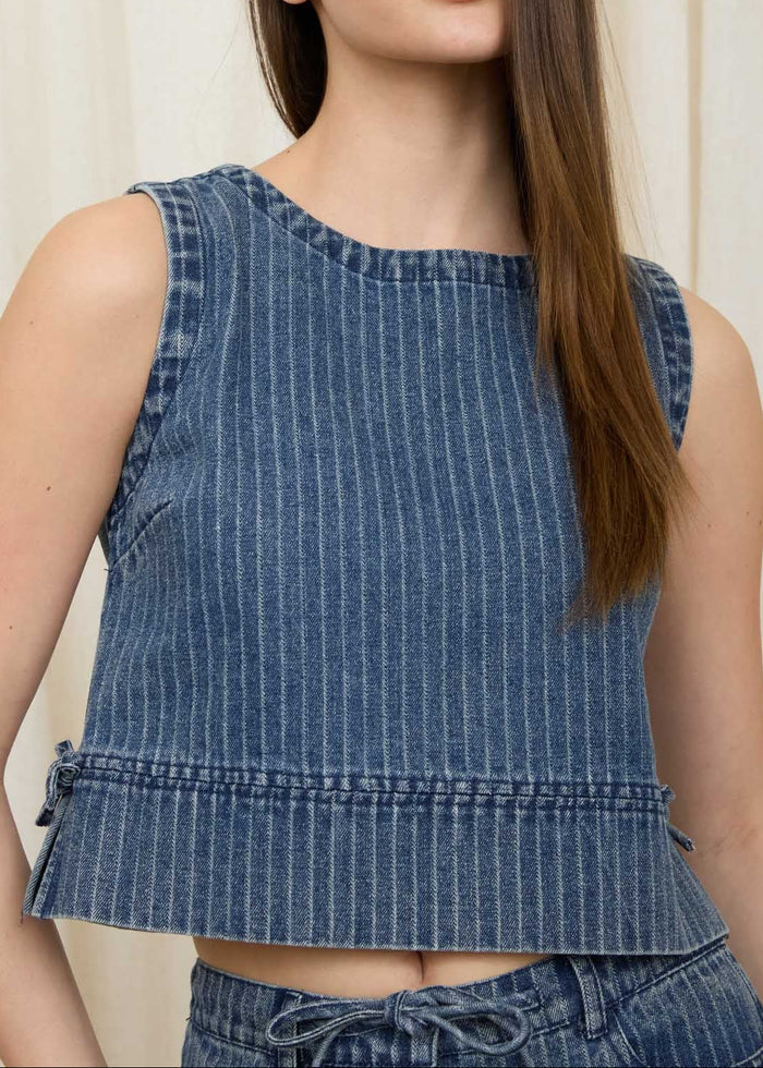 Woman wearing a blue denim sleeveless top and shorts against a white curtain background