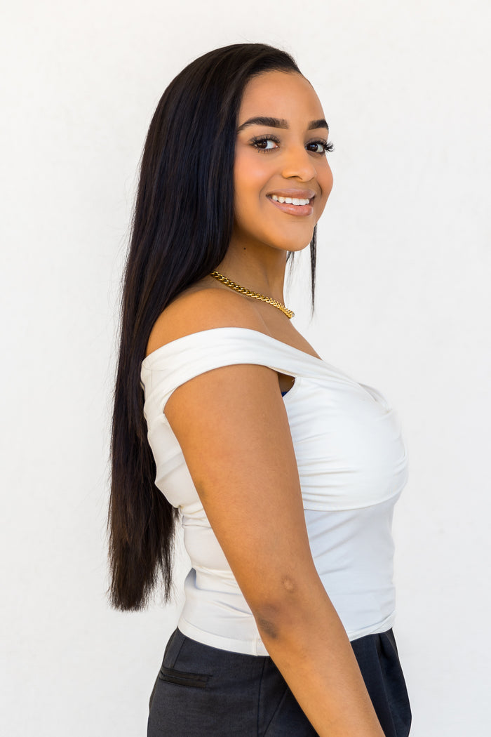 White off-the-shoulder top with ruched fabric detail and short sleeves on model wearing black pants.