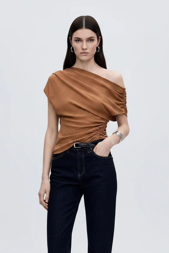 Woman wearing tan one-shoulder top with ruched side detail and dark denim jeans.