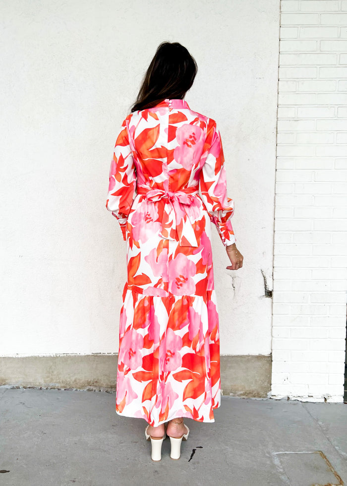 Woman wearing long-sleeved maxi dress with red, pink, and orange floral print on white background, tied at waist.