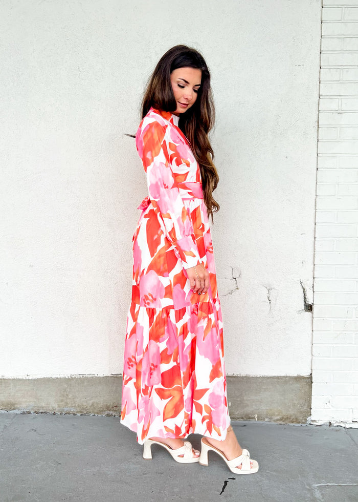Woman wearing a long-sleeved maxi dress with red and pink floral print on white background, paired with white sandals.