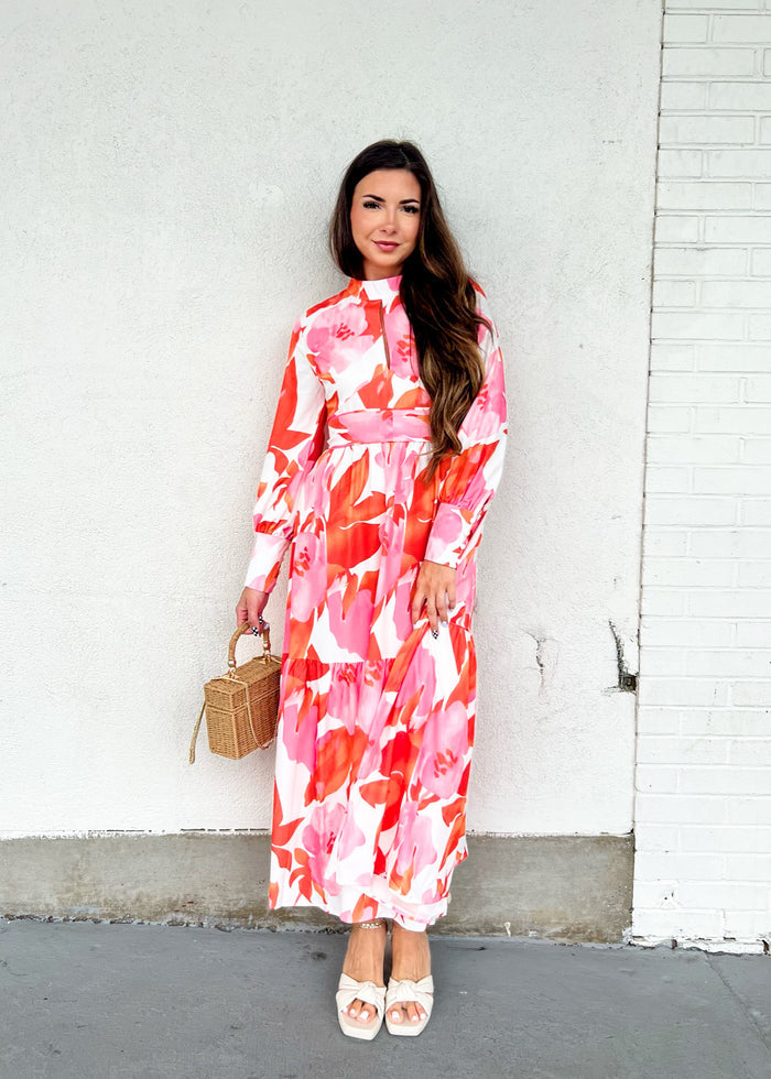 Woman wearing long-sleeve floral maxi dress in pink, red, and white with collar neckline and belted waist.