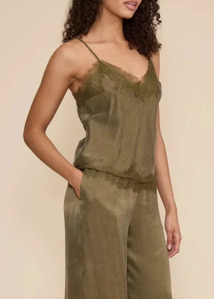 A woman modeling a sage green sleeveless jumpsuit with thin straps and wide-leg harem-style pants, standing against a plain beige studio background.
