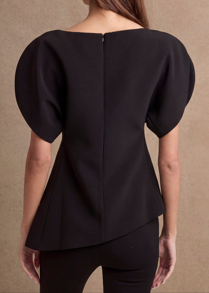 A woman viewed from the back wearing a black peplum-style top with short puff sleeves and a high-low hem, against a warm beige textured backdrop.