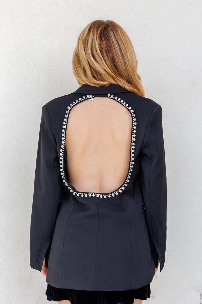 Black blazer with open back cutout outlined in rhinestones, long sleeves, worn over black skirt.